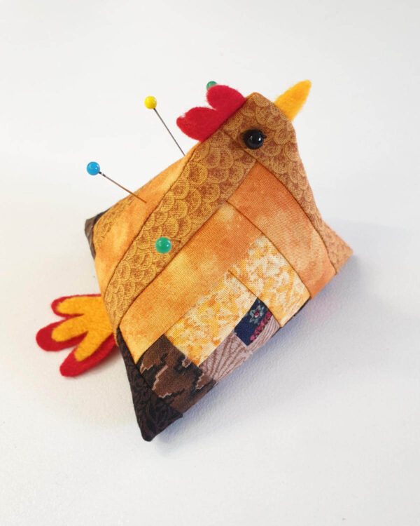 The photo shows a Handmade pin cushion chicken crafted from log cabin quilt blocks, with decorative pins inserted, set against a plain white background.