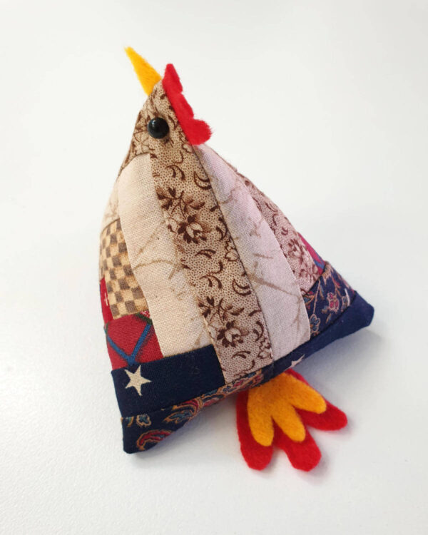 Handmade plush toy shaped like a chicken, adorned with log cabin quilt blocks in various patterns and colors, featuring a red felt comb and beak, along with yellow and red felt feet.