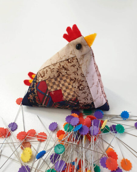 The handmade pin cushion chicken, featuring log cabin quilt blocks, sits on a white surface surrounded by colorful flower-head pins, ideal for piecing together quilt blocks.