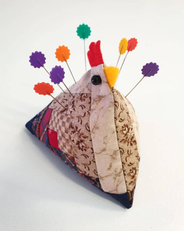 Handmade "Pin Cushion Chicken" with log cabin quilt blocks and colorful pins on its back, elegantly arranged on a pristine white surface.