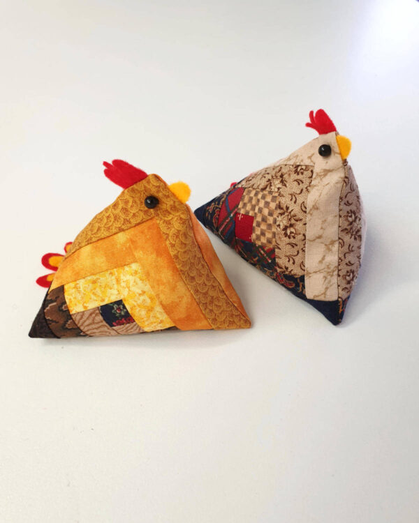 Two handmade pin cushion chickens, adorned with patchwork designs and featuring a red felt comb and yellow beak, rest on a white surface. These charming creations echo the rustic charm of log cabin quilt blocks.